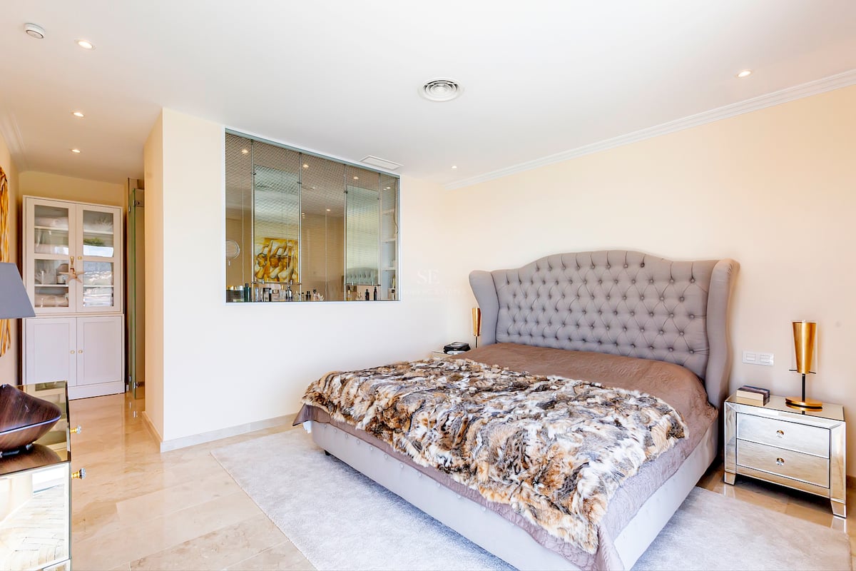 Chambre spacieuse avec tête de lit capitonnée grise, sol en marbre, tables de chevet en miroir et rangements.