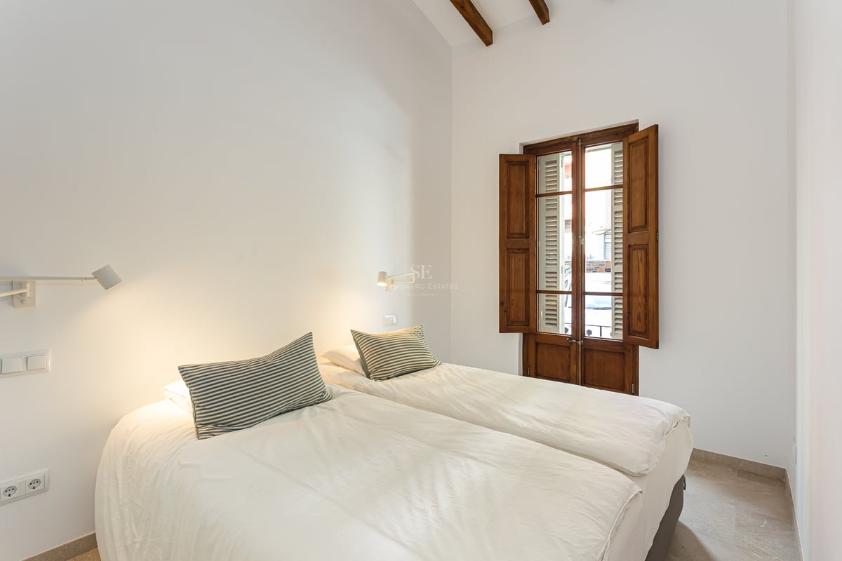 Chambre minimaliste blanche avec deux lits, hauts plafonds avec poutres et volets traditionnels en bois.