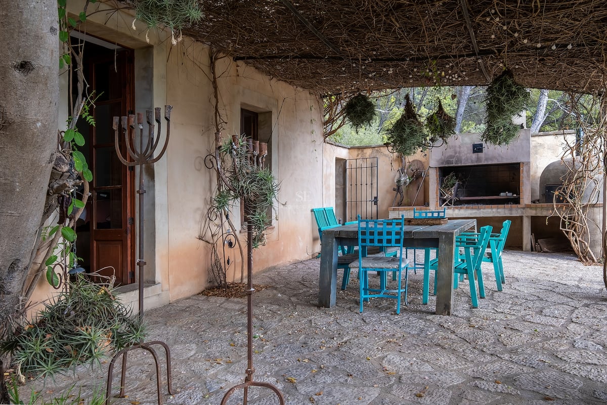 Terrasse en pierre avec table en bois, chaises bleu turquoise, toit en brande et un coin barbecue à l'arrière-plan.