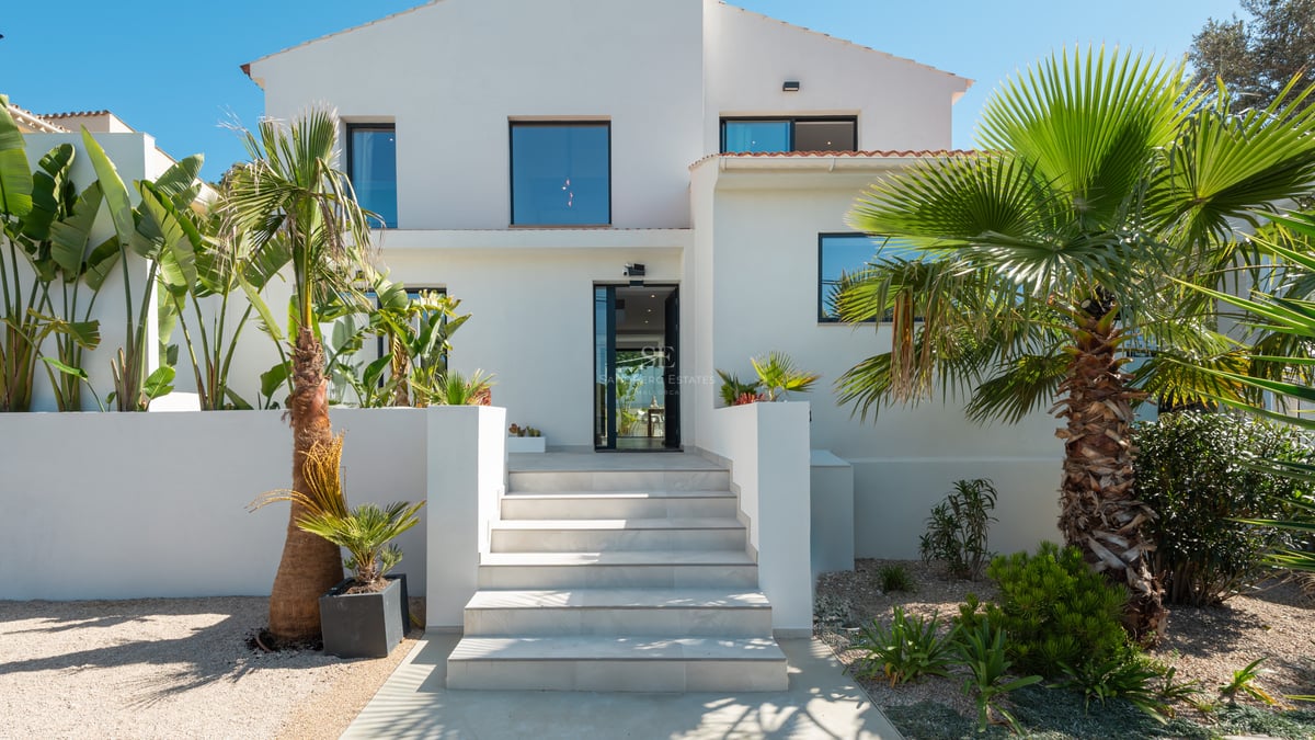 Extérieur d'une villa blanche avec un escalier en pierre claire, une porte vitrée et une végétation tropicale.