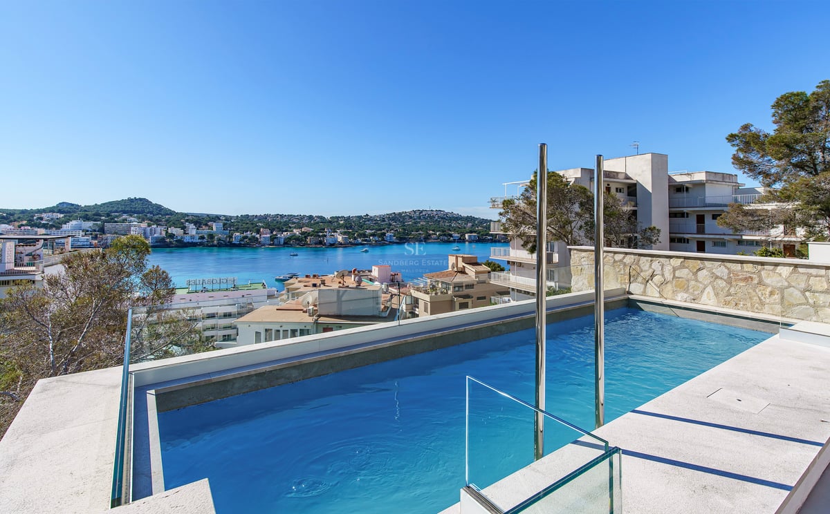 Piscine moderne avec garde-corps en verre surplombant une baie méditerranéenne et une ville côtière sous un ciel bleu.