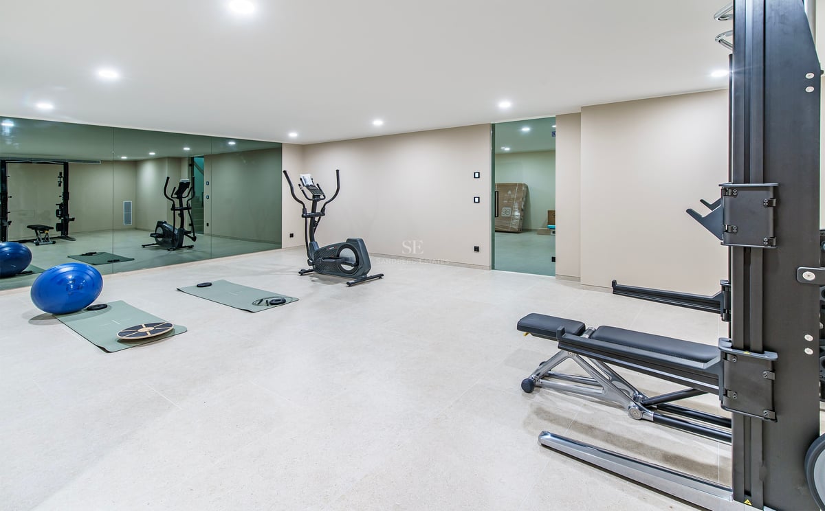 Salle de sport avec vélo elliptique, banc de musculation et grands miroirs sous des plafonniers brillants.