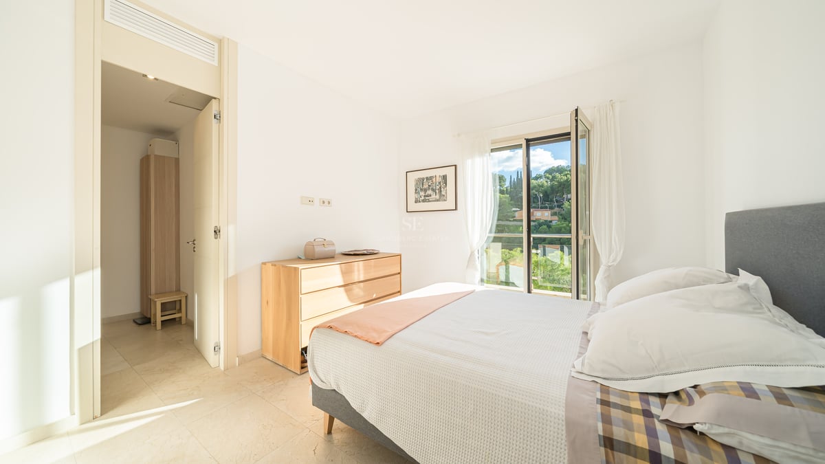 Chambre lumineuse avec lit, commode en bois clair et porte de balcon ouverte donnant sur la verdure.