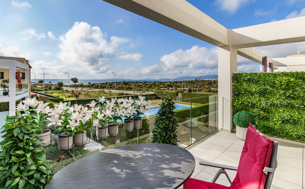 Terrasse de luxe avec table ronde, chaise rouge et garde-corps en verre surplombant jardin, piscine et mer.