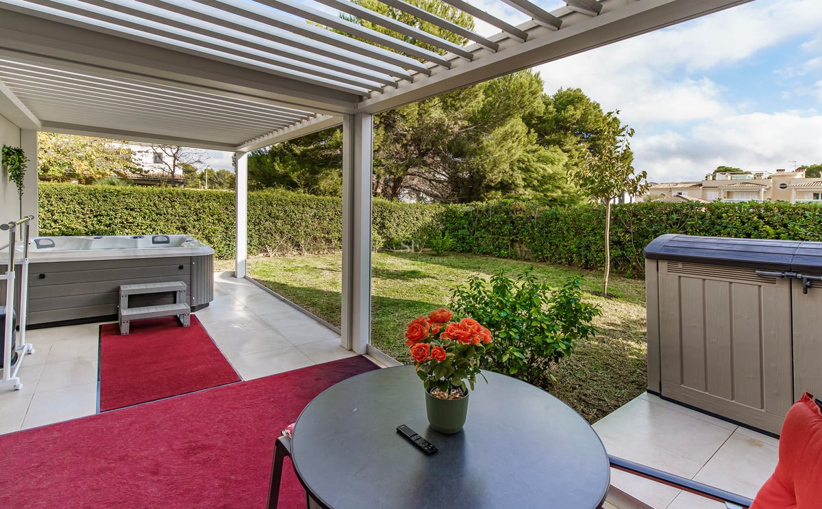 Terrasse avec pergola blanche, jacuzzi gris, tapis rouges et vue sur un jardin verdoyant avec haies.