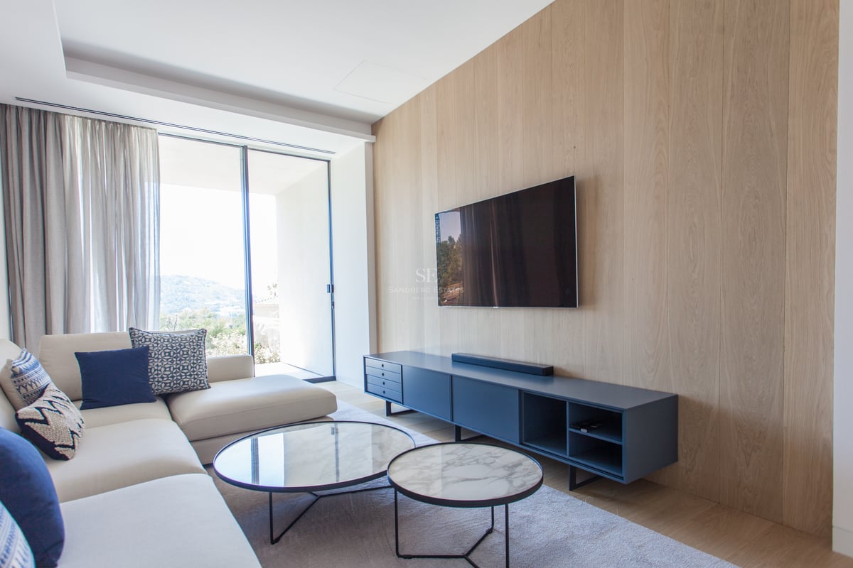 Salon moderne avec mur en chêne, télévision murale, tables en marbre et grandes baies vitrées vers les collines.