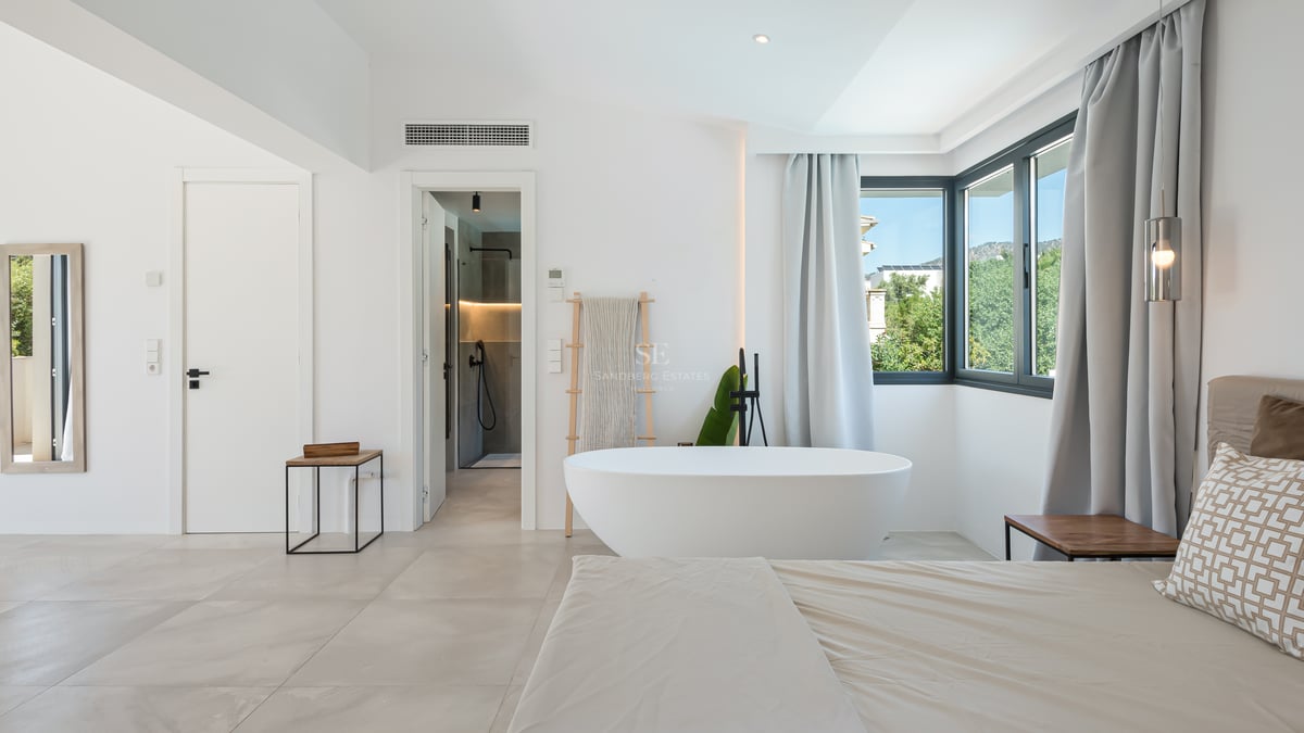 Grande chambre blanche avec baignoire îlot ovale, rideaux gris et salle de bain attenante visible à l'arrière-plan.