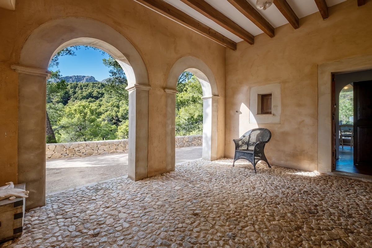 Terrasse rustique avec arches, poutres en bois, sol en galets et vue sur la forêt.
