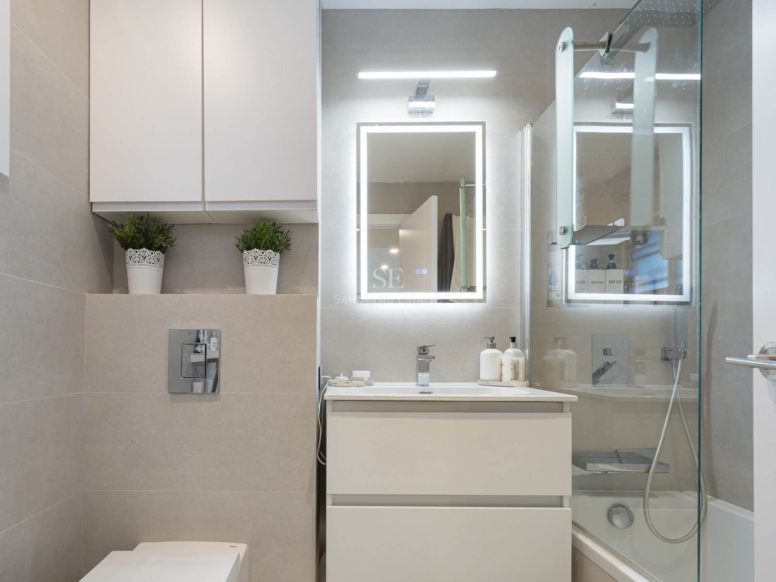 Salle de bain moderne avec vanité blanche, miroir LED, paroi en verre et carrelage beige grand format.