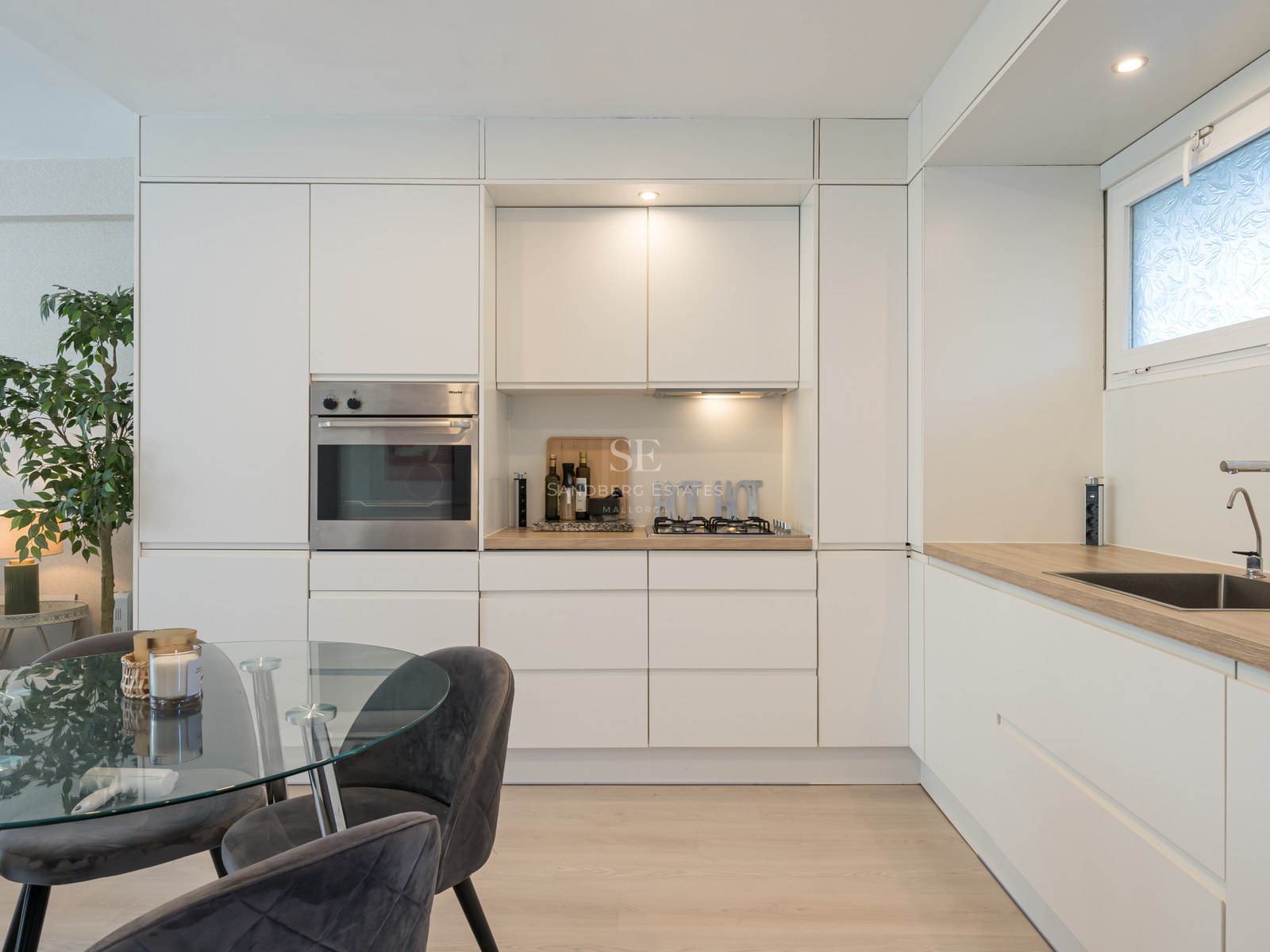 Cuisine minimaliste blanche avec four Miele, plan de travail effet bois et table à manger en verre.
