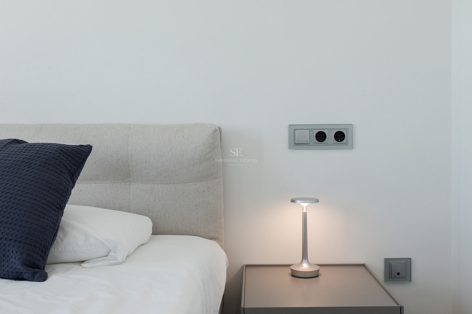 Close-up of a bed with grey headboard, nightstand with a designer lamp, and modern electrical sockets on a white wall.