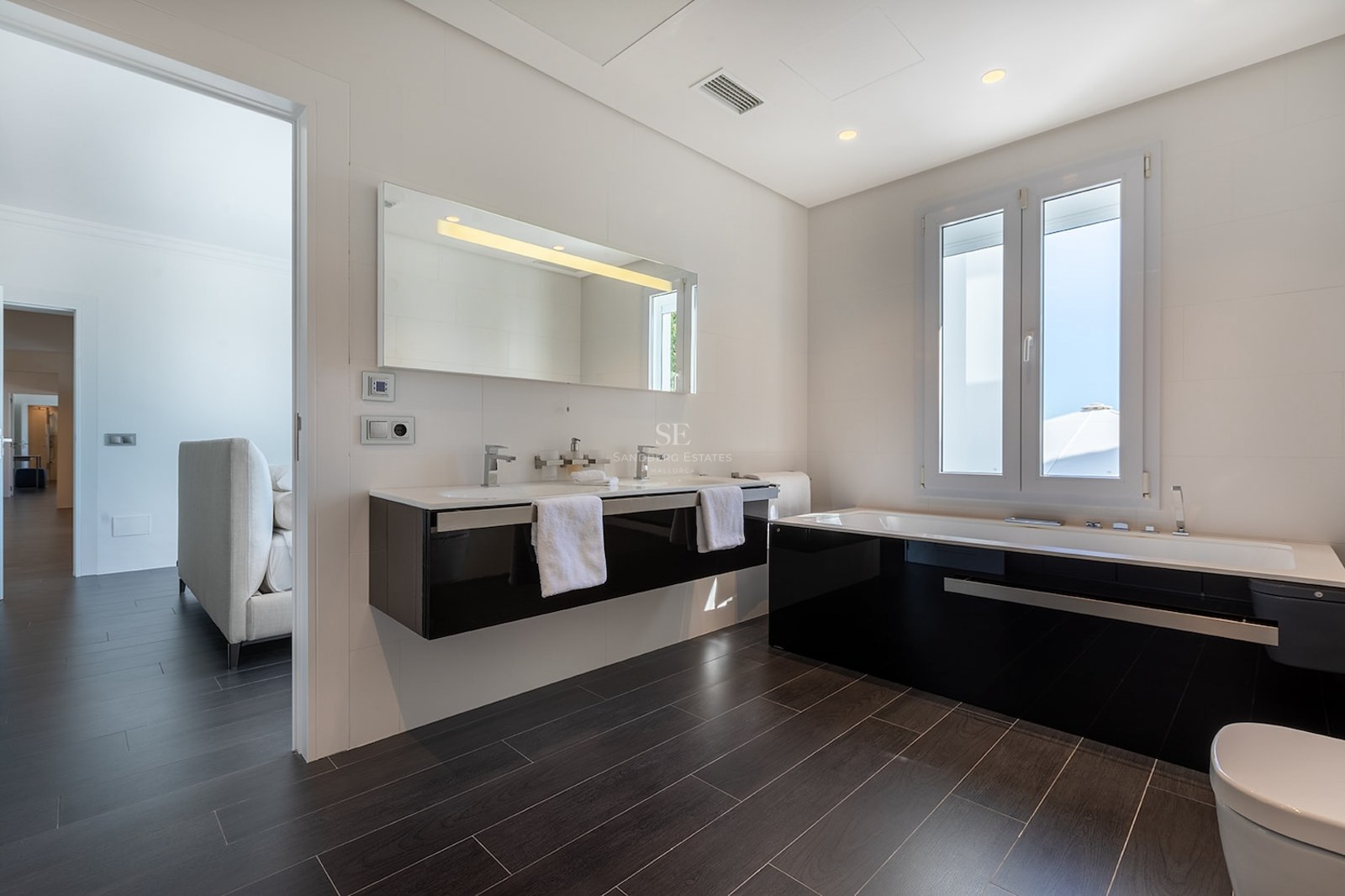 Modern bathroom with double vanity, black bathtub, and dark flooring next to a bright bedroom.