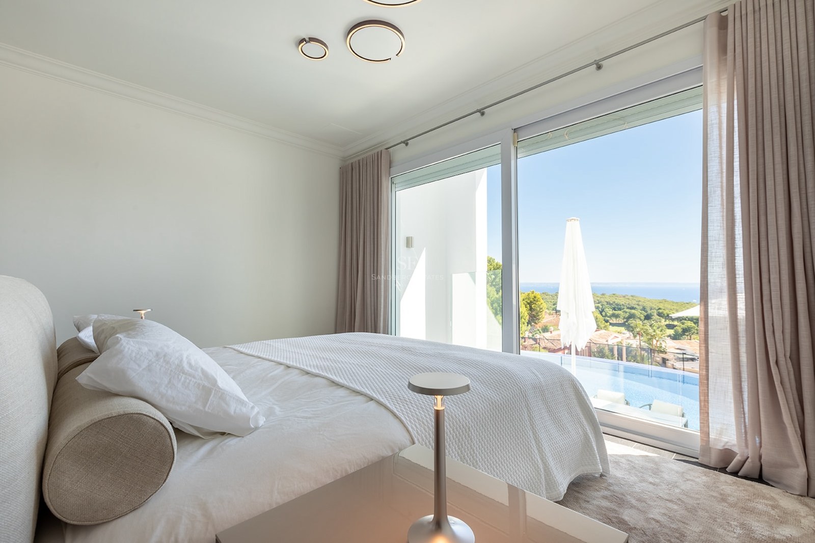 Modern bedroom with white bedding, large windows overlooking a private infinity pool and distant sea view.