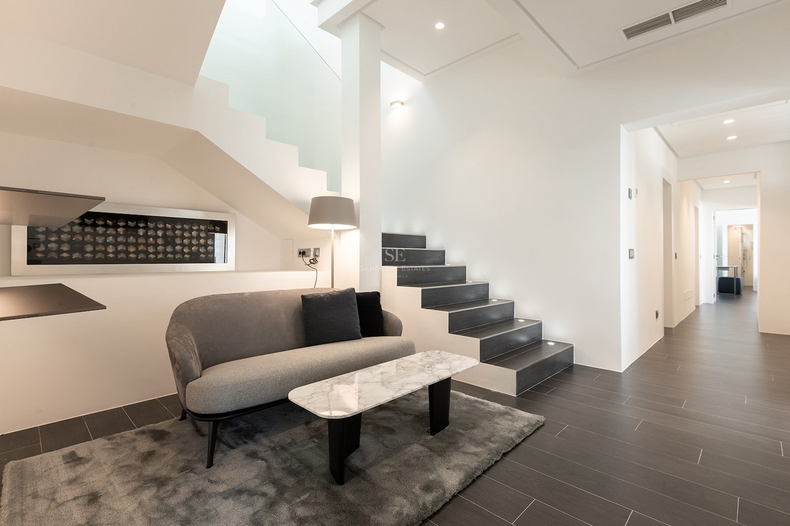 Modern lounge area featuring a grey sofa, marble table, and a contemporary dark-tread staircase with integrated lighting.
