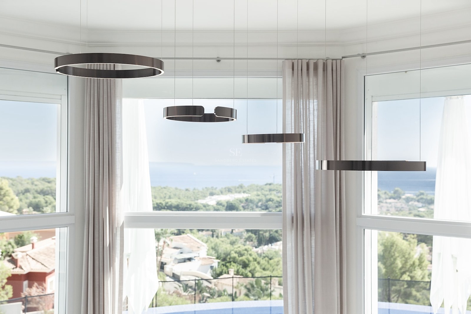Four bronze ring-shaped pendant lights hanging in front of a large bay window overlooking a coastal landscape and the sea.