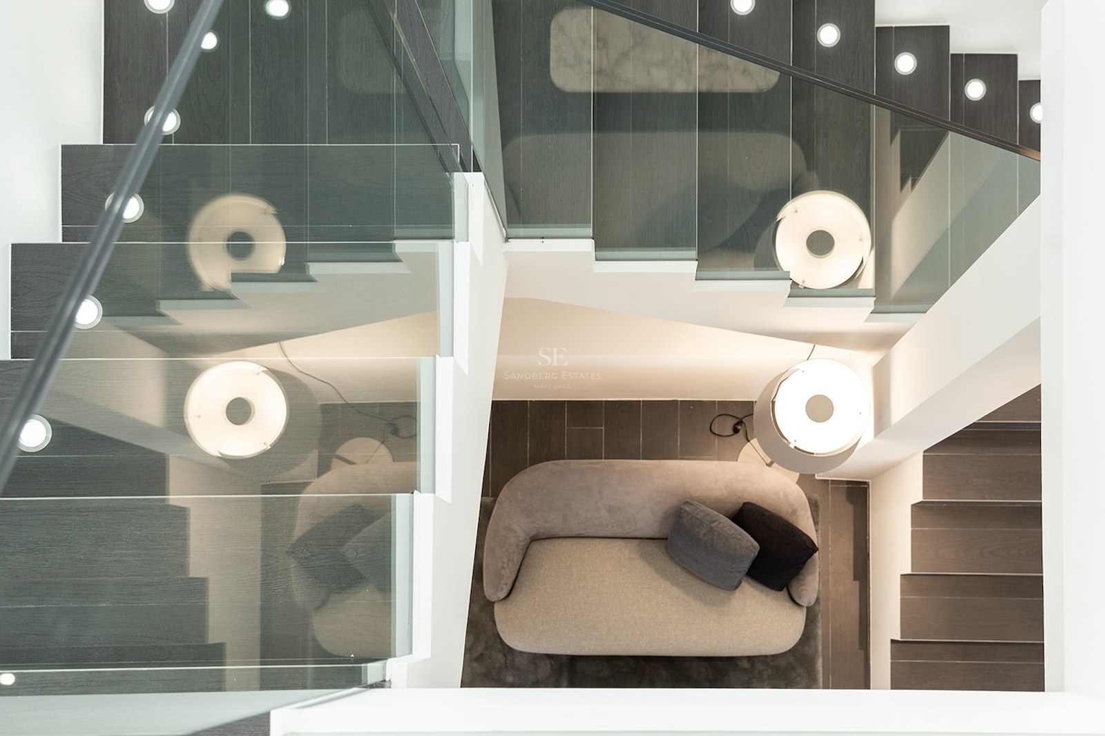 Overhead view of a staircase with glass railings looking down onto a grey sofa and a designer lamp.
