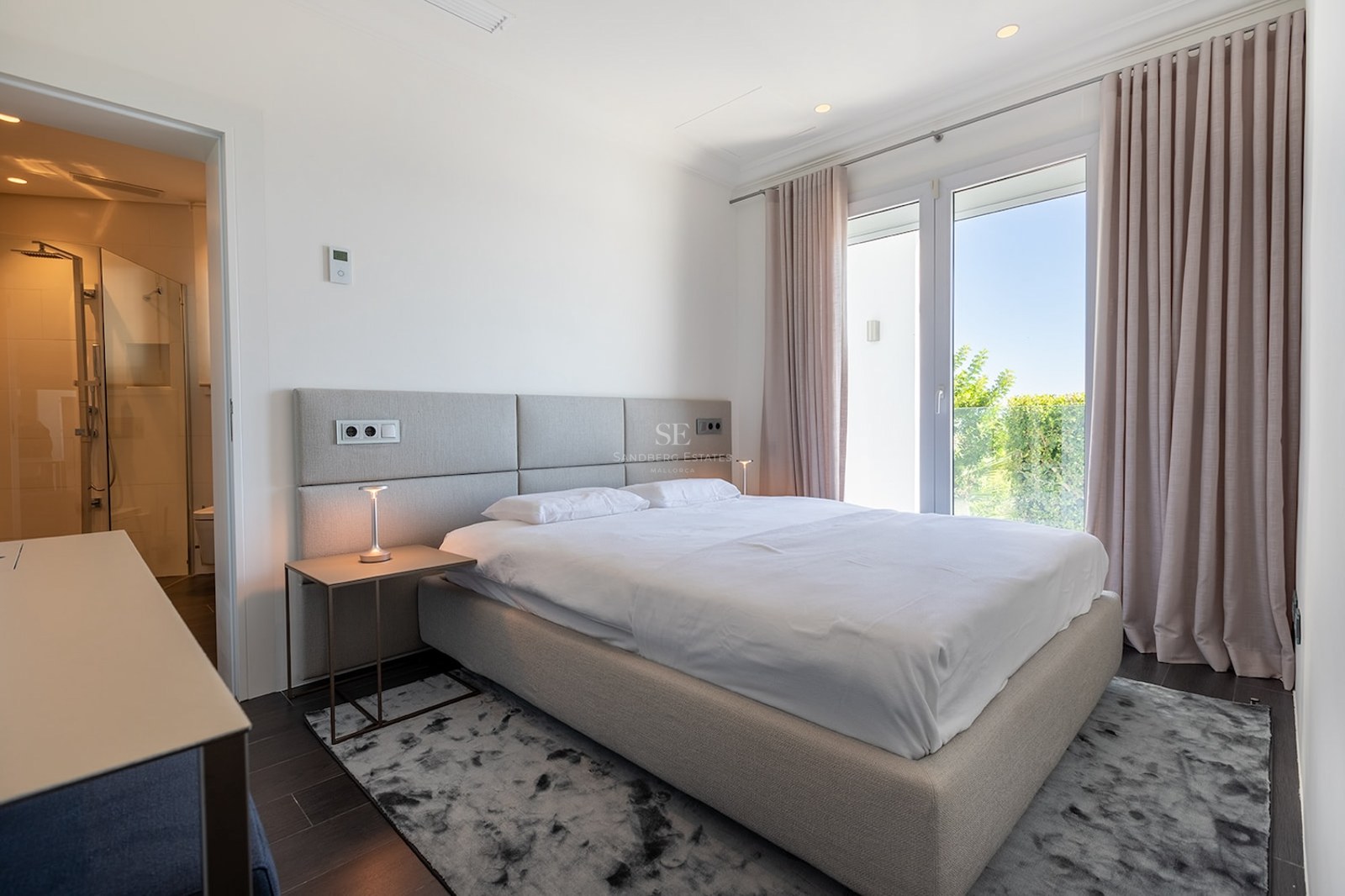 Bright master bedroom with a grey upholstered bed, white linens, ensuite bathroom, and large windows looking onto greenery.