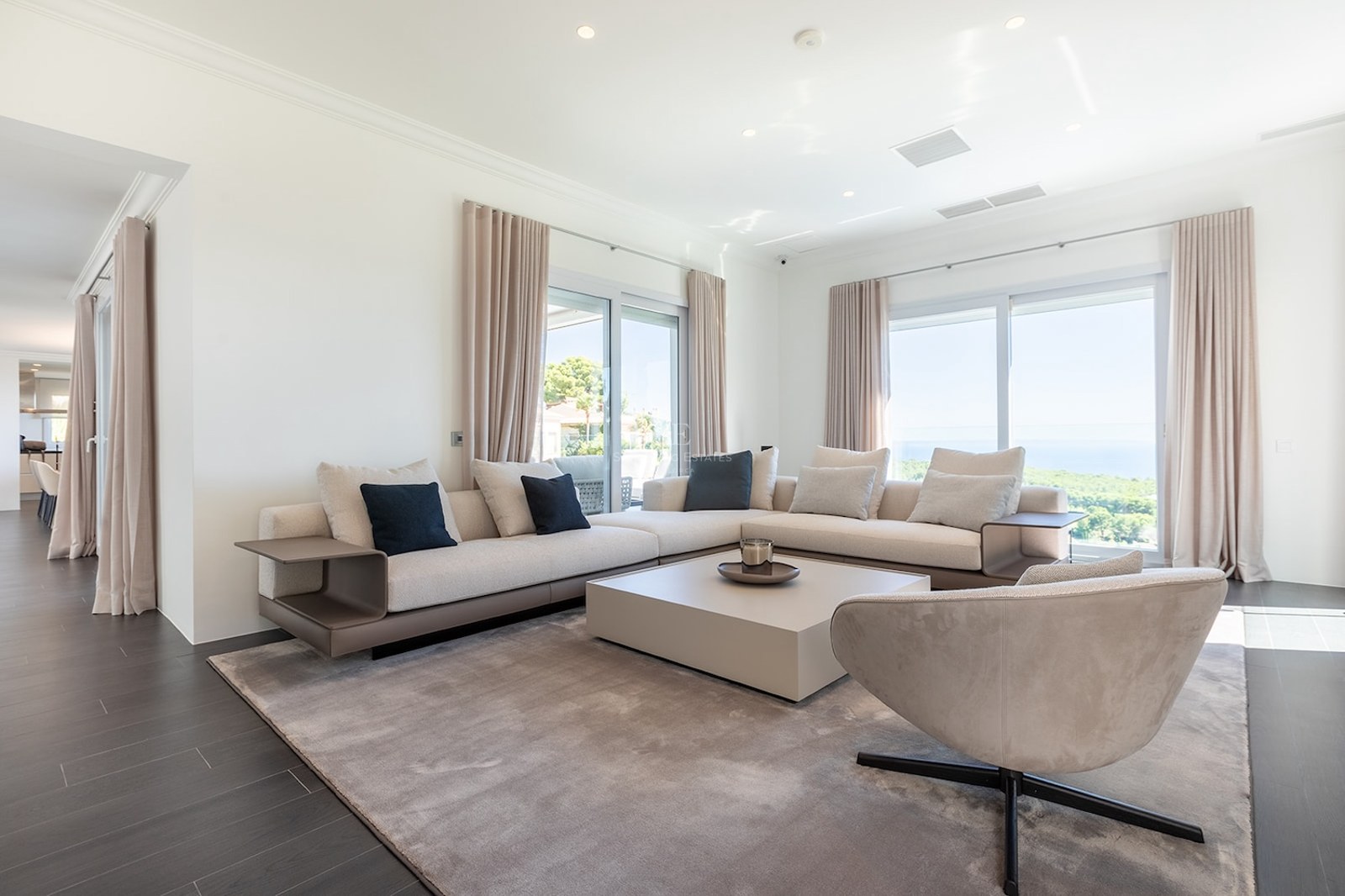Spacious white living room with beige sectional sofa, designer armchair, and large windows overlooking the ocean.