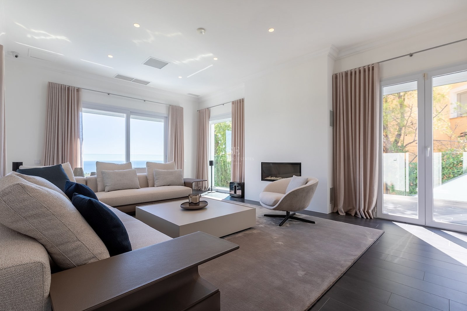 Spacious living room with cream sofas, built-in fireplace, dark flooring, and large windows overlooking the sea.