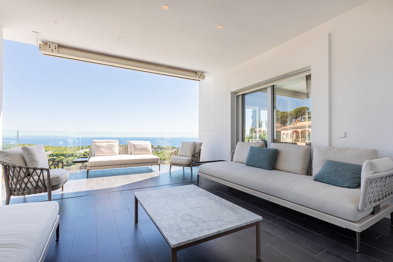 Modern covered terrace with white sofas, a marble coffee table, and a glass railing overlooking the Mediterranean sea.