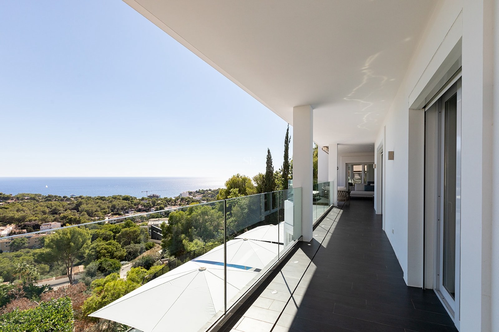 Spacious white balcony with glass railing overlooking a Mediterranean forest and the blue ocean.