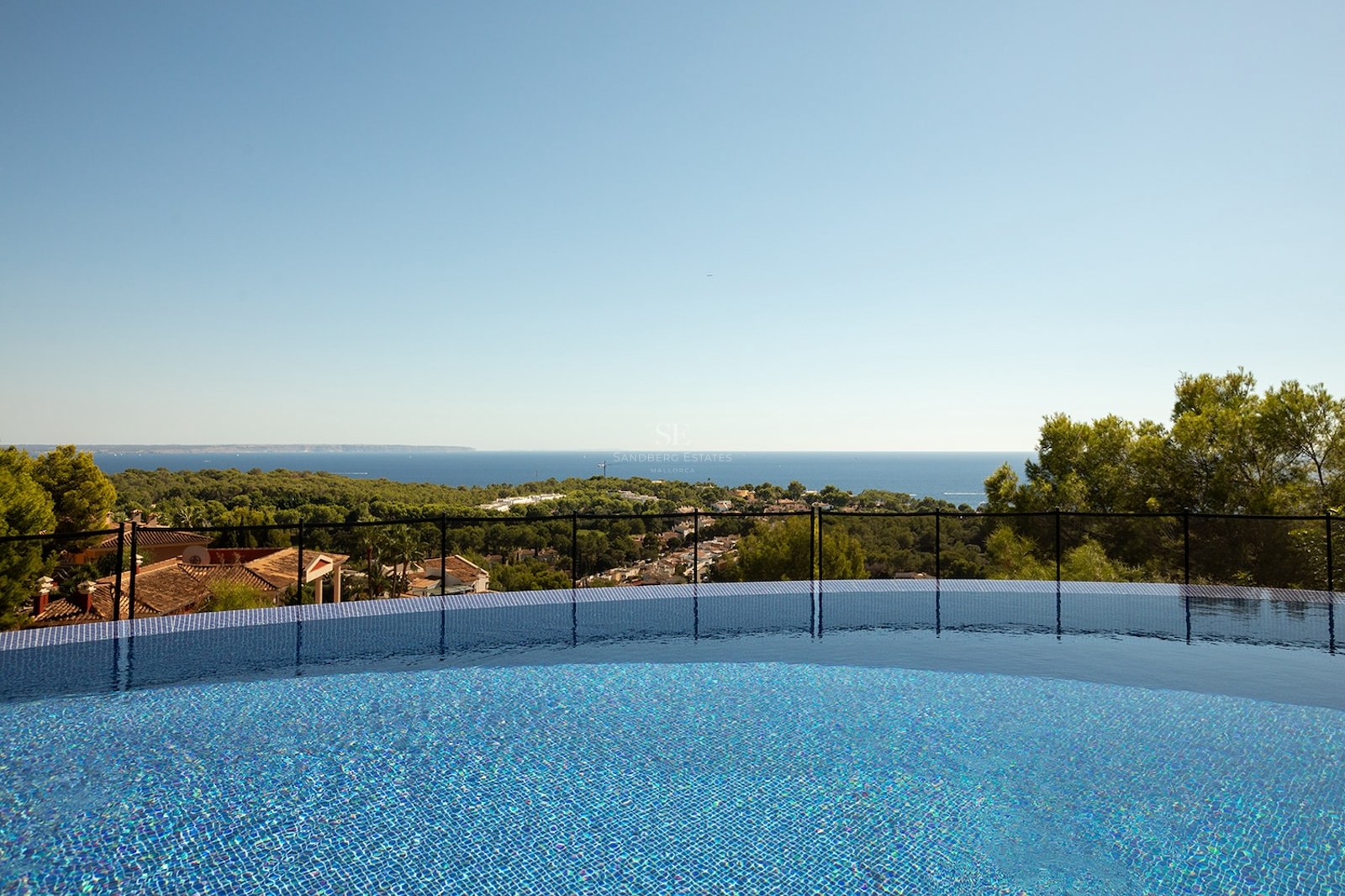 View from a blue mosaic infinity pool looking out over green trees towards the Mediterranean sea under a clear sky.