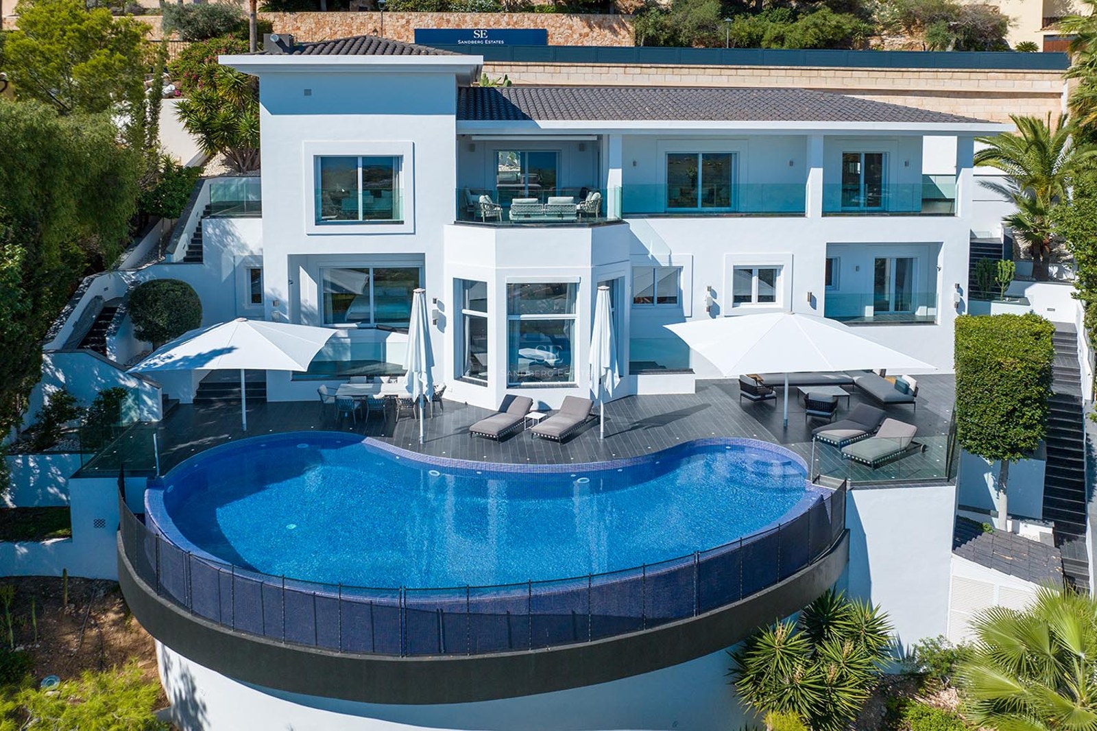 Elevated view of a modern white villa featuring a large blue semicircular infinity pool and furnished terraces.