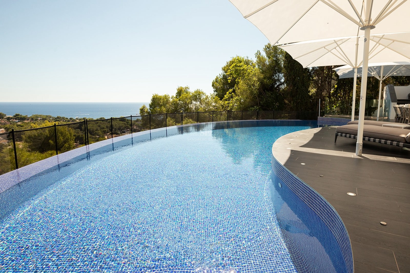 Curved infinity pool with blue mosaic tiles overlooking a lush green landscape and the blue ocean under a clear sky.