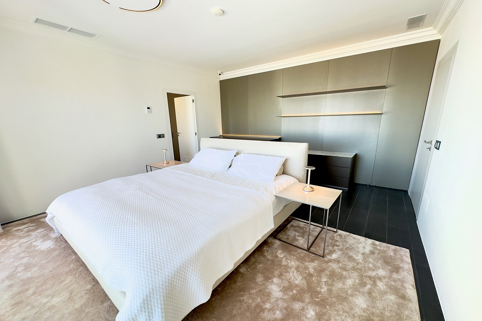 Modern bedroom featuring a large white bed, minimalist nightstands, a taupe feature wall, and a plush beige rug.