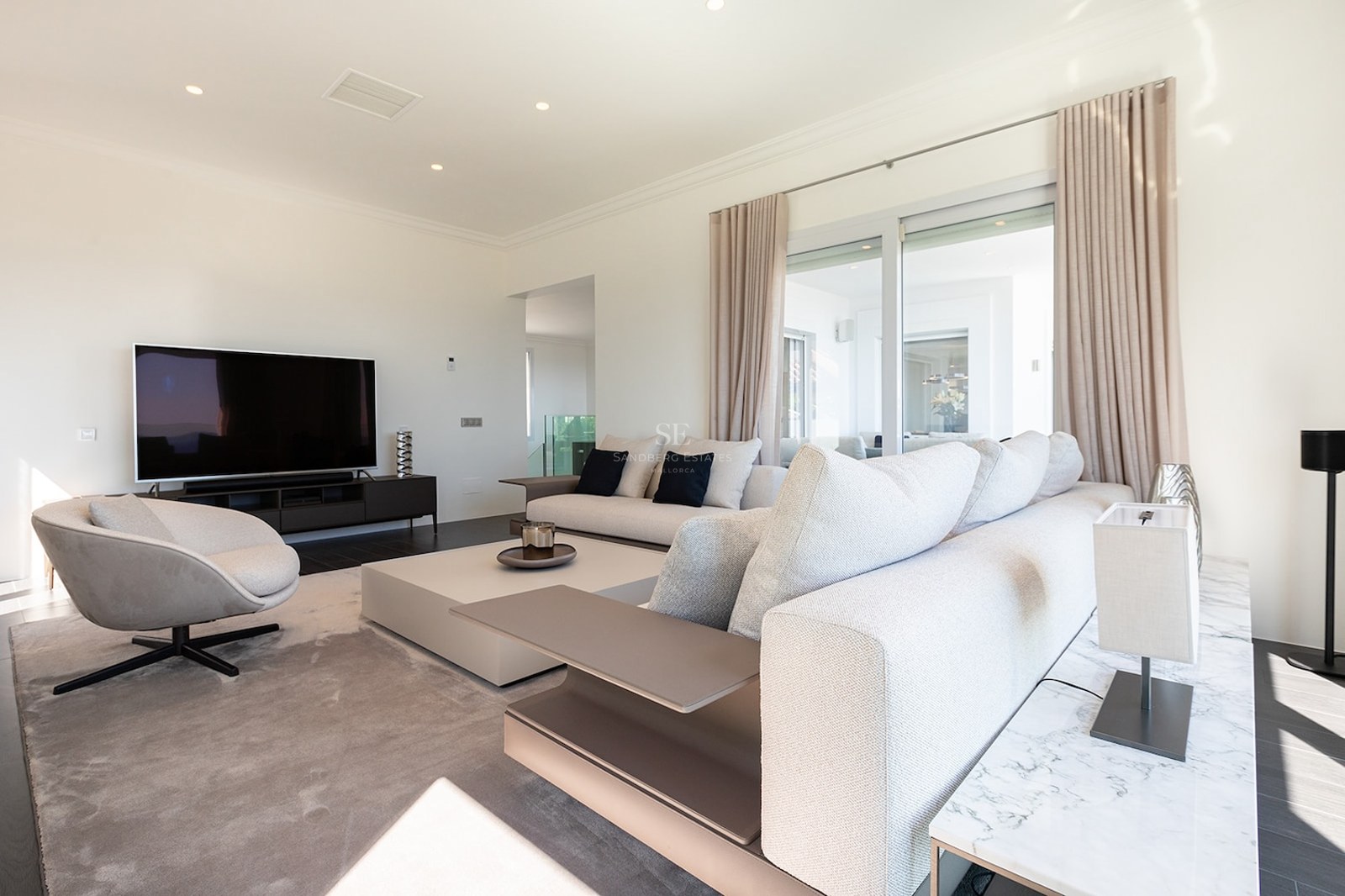 Contemporary living room featuring a large beige sofa, a designer armchair, and a flat-screen TV in a bright space.