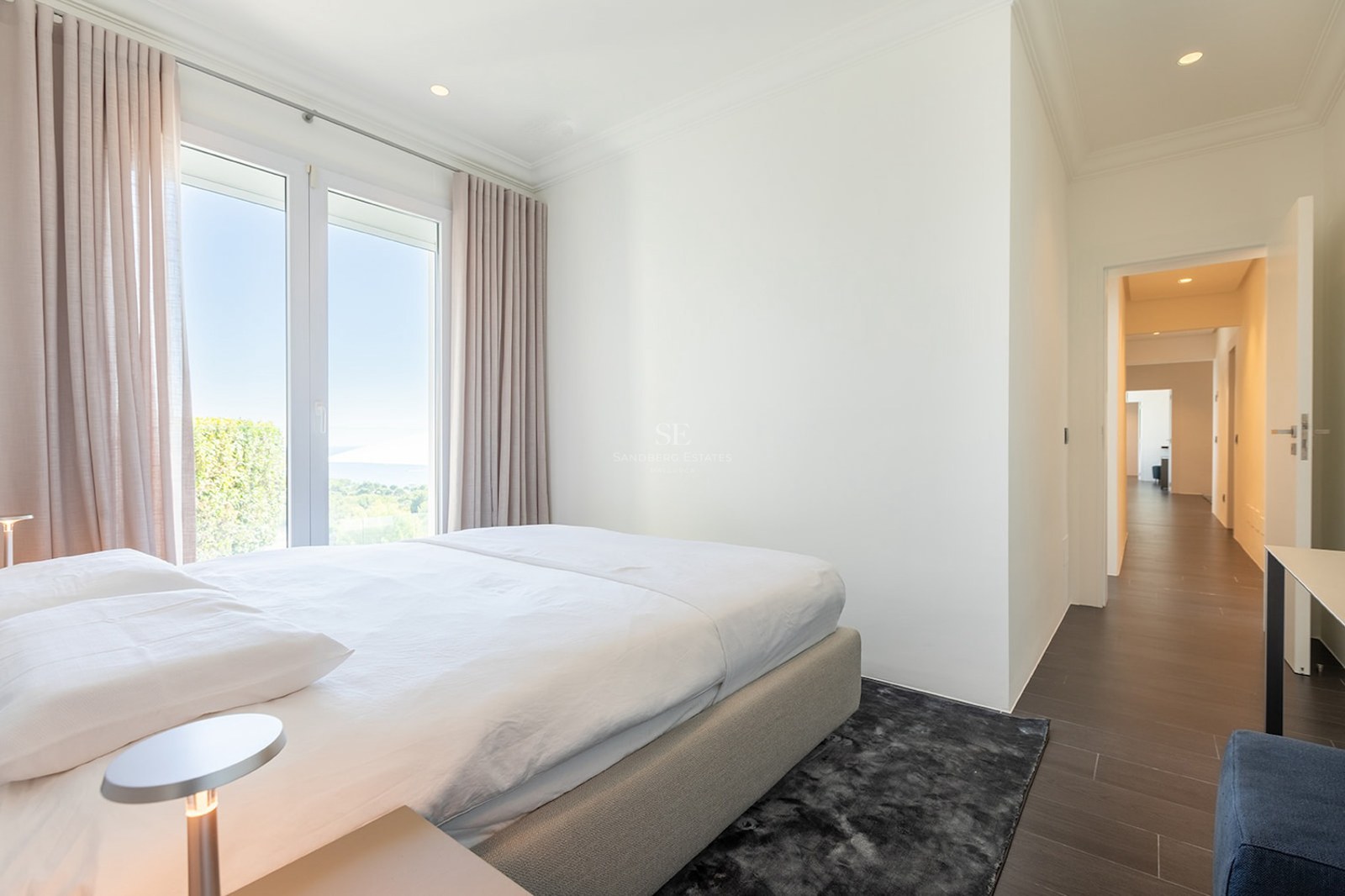 Bright bedroom featuring a white bed, dark rug, and large window overlooking the sea and lush greenery.