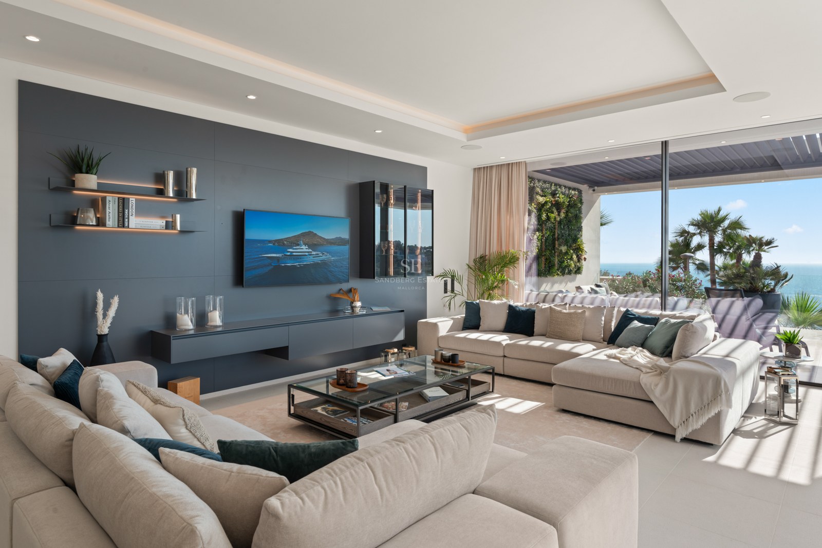 Bright living room with large beige sofas, charcoal accent wall, TV, and panoramic glass doors opening to a sea view terrace.