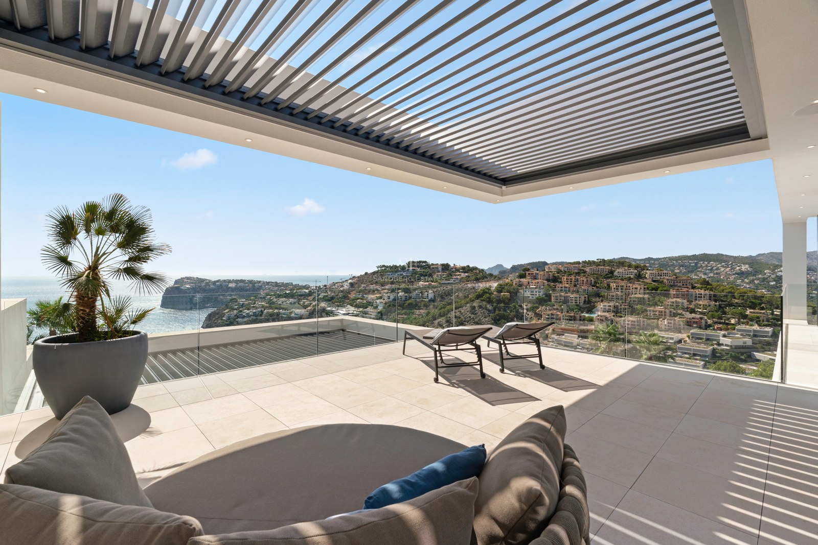 Modern terrace with stone tiling, sun loungers, a louvered pergola, and stunning views of the sea and coastal hills.