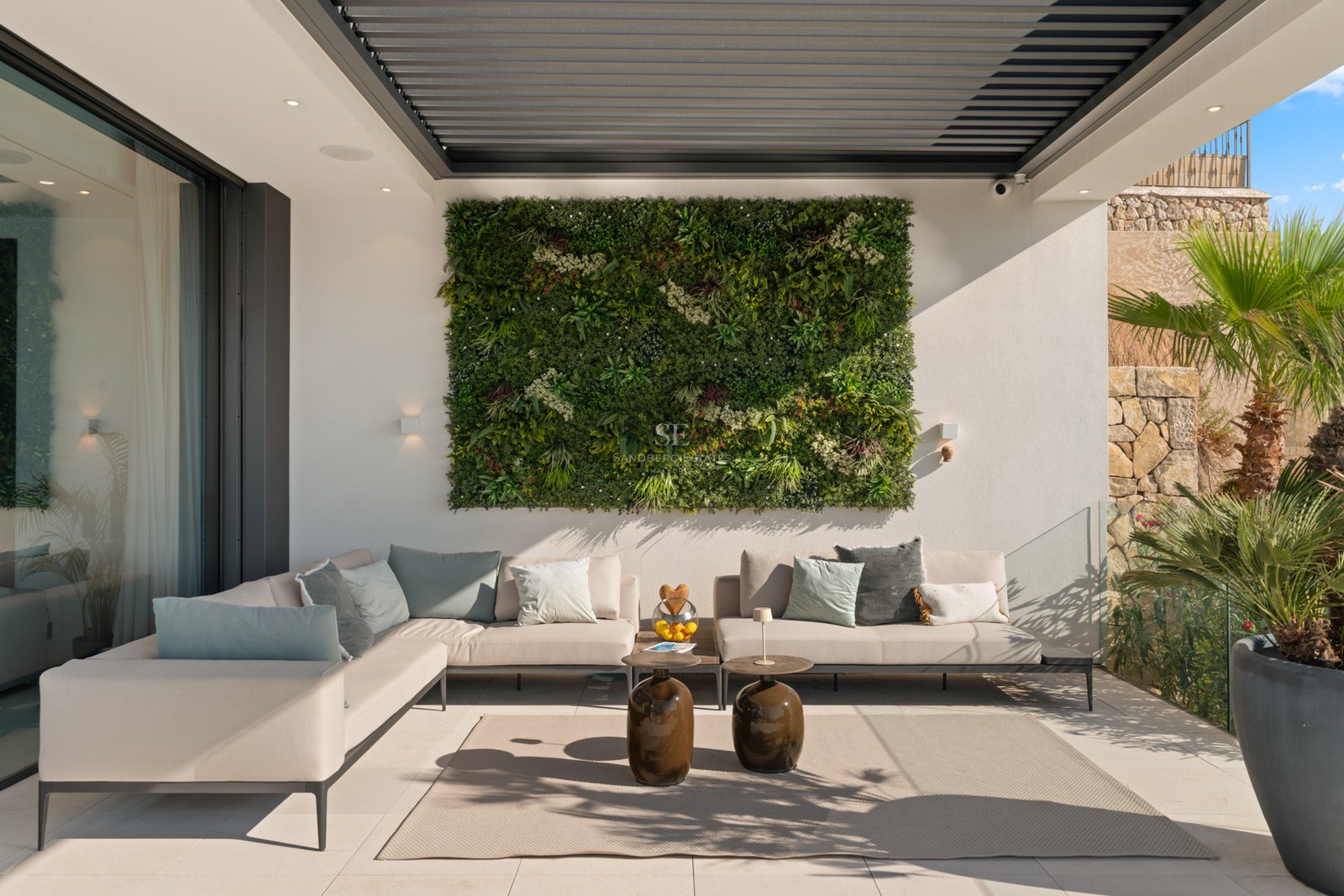 A contemporary outdoor terrace featuring a large beige corner sofa, a lush green vertical garden wall, and a louvered pergola.