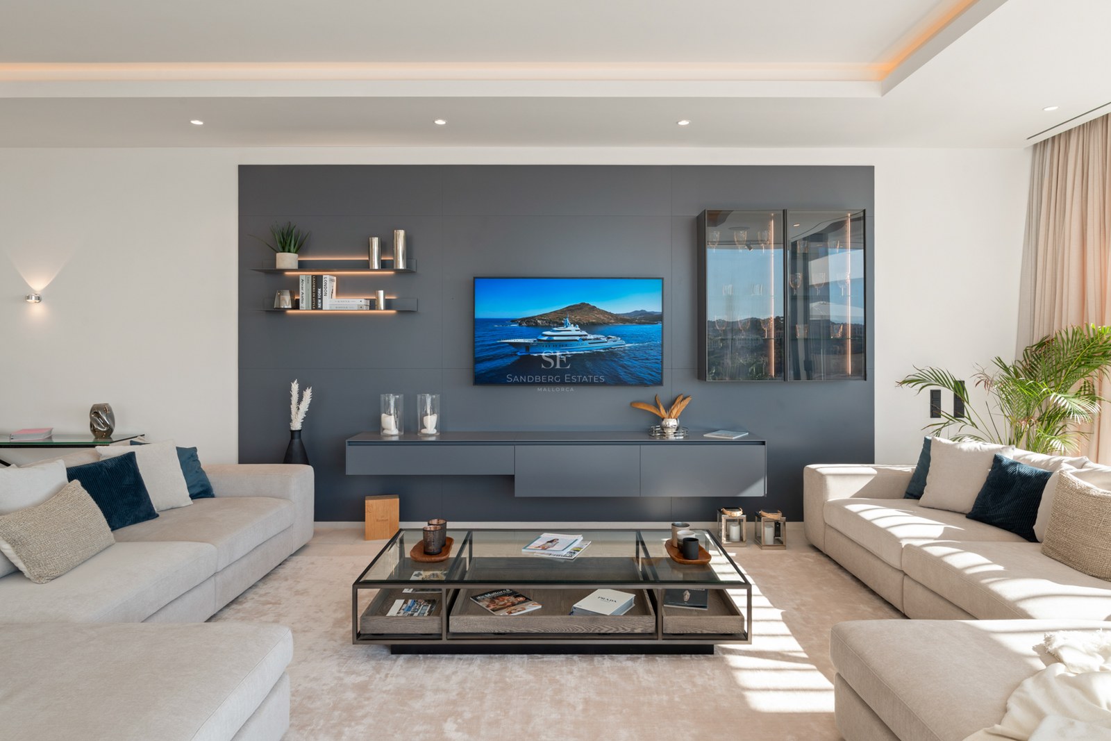 A bright, modern living room with cream sofas, a large grey media wall with TV, and a glass coffee table.