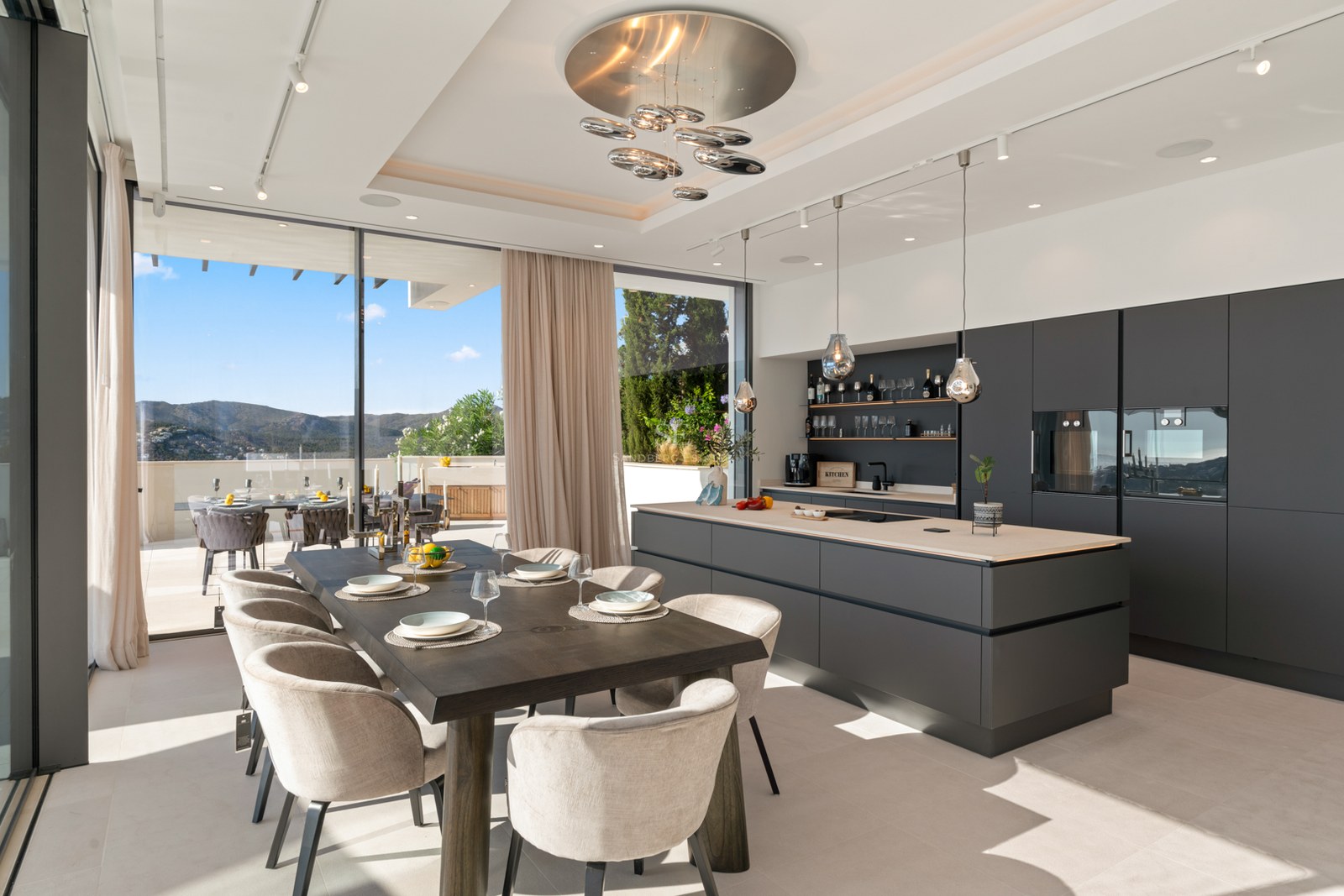 Modern kitchen with large grey island and wooden dining table next to floor-to-ceiling windows with mountain views.