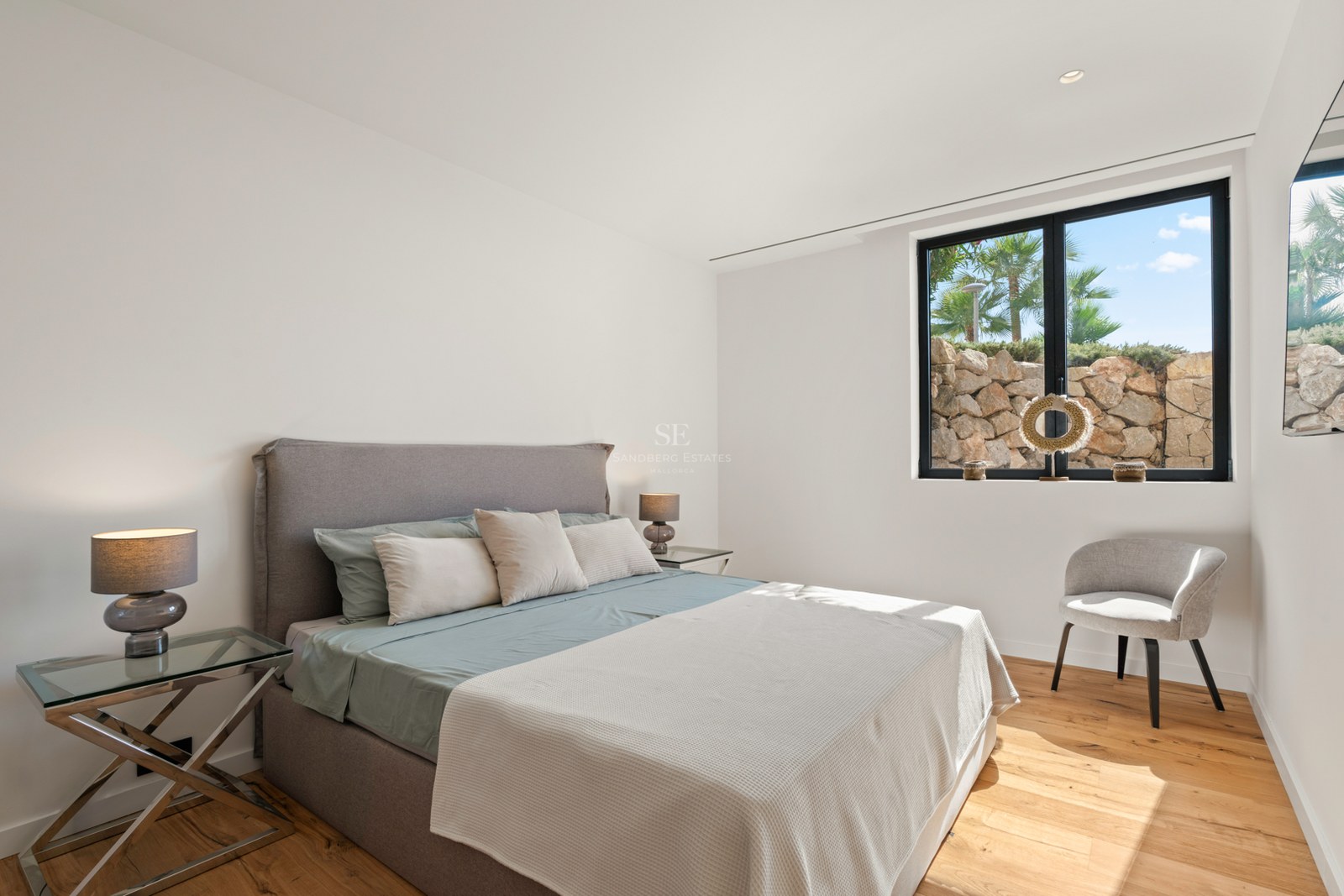 Bright modern bedroom with a gray upholstered bed, glass nightstands, wooden floors, and a large window.