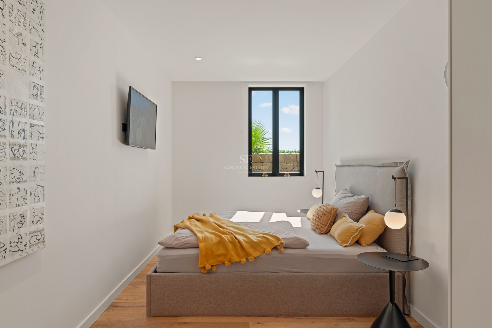 Bright bedroom featuring a taupe upholstered bed, yellow pom-pom blanket, minimalist lamps, and a scenic window view.