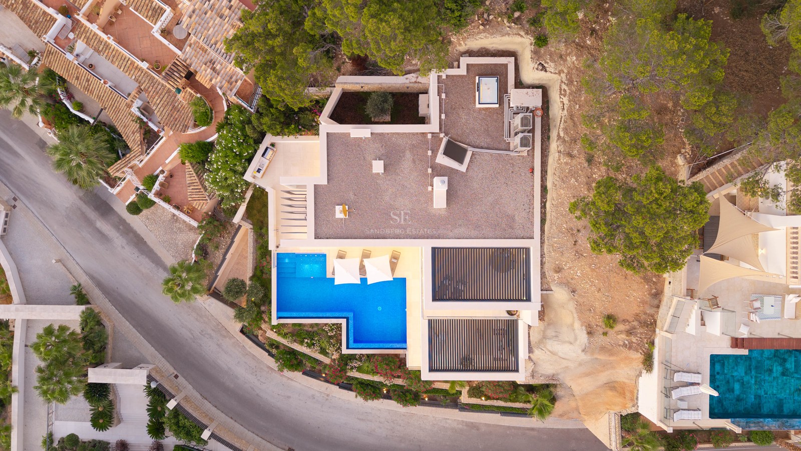 Flygvy uppifrån av en modern vit villa med en klarblå L-formad pool och solstolar.