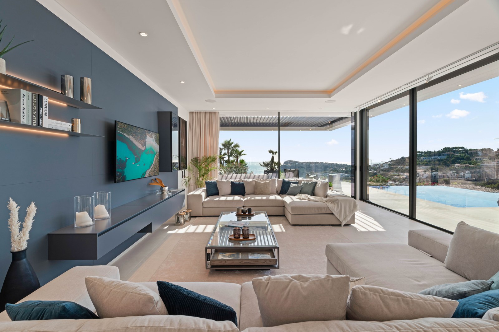 Spacious modern living room with beige sofas, a blue accent wall, and large windows overlooking the sea and an infinity pool.