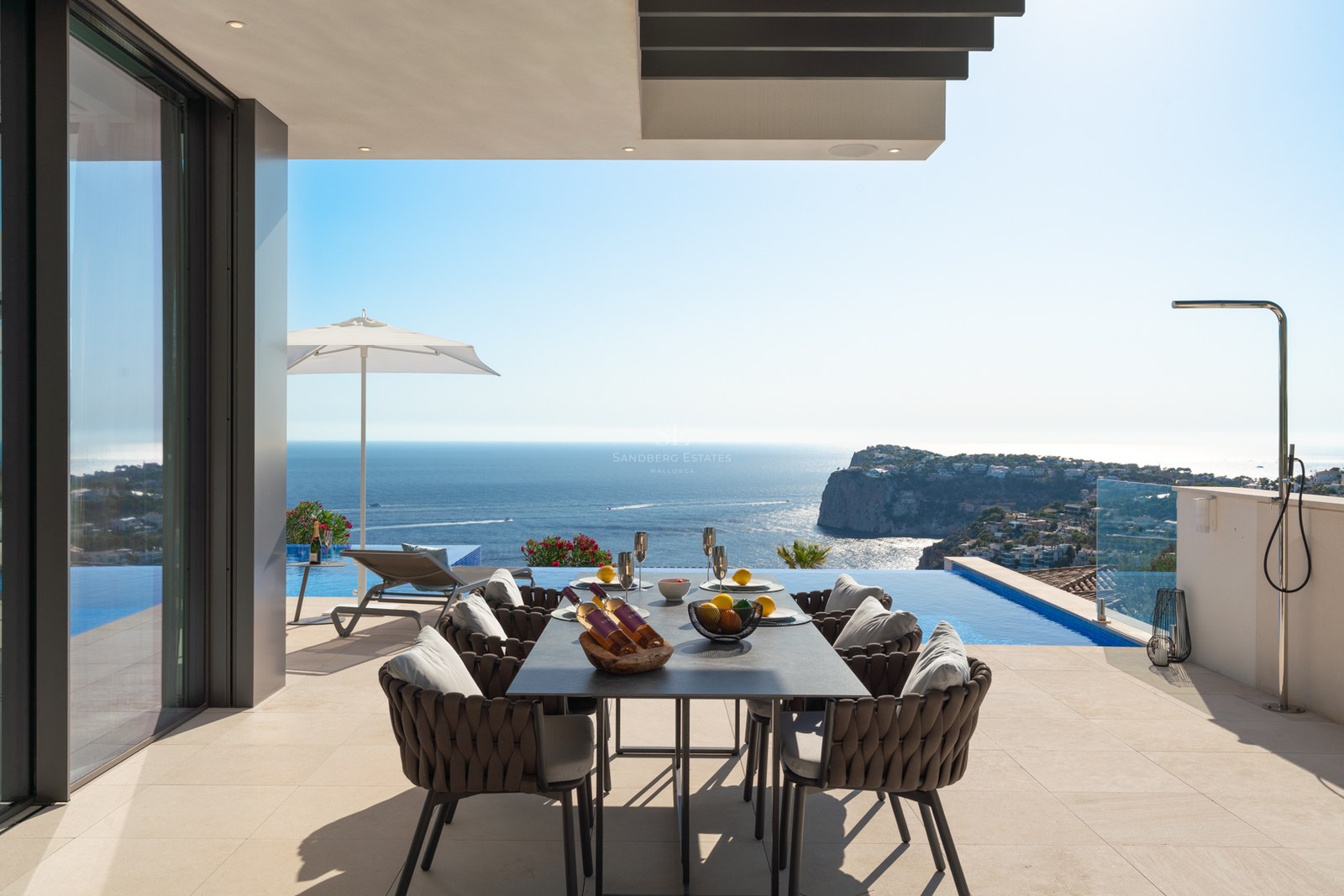 Modern outdoor terrace with a dining table overlooking an infinity pool and a vast blue ocean with coastal cliffs.