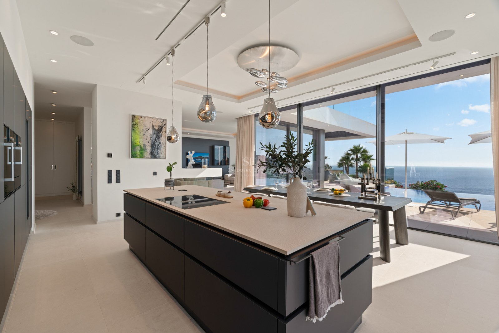 Modern kitchen with a large dark island, designer glass pendants, and floor-to-ceiling windows overlooking the ocean.