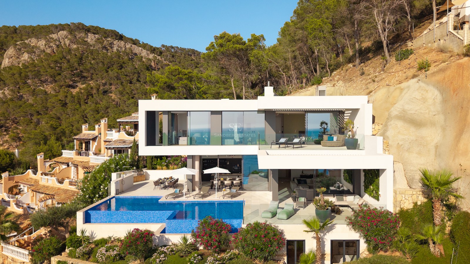 Multi-level modern white villa on a hillside featuring a large infinity pool, lounge terraces, and lush greenery.
