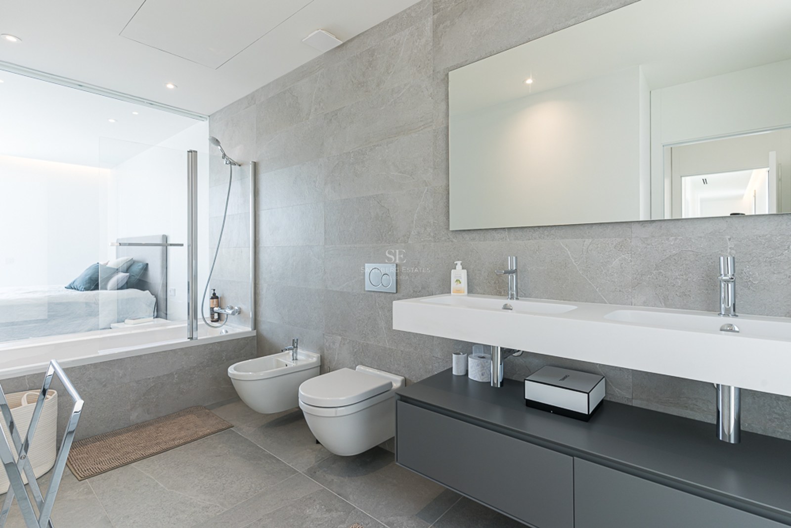 Modern bathroom featuring grey stone tiles, double vanity, wall-hung toilet, and a glass wall looking into a bedroom.