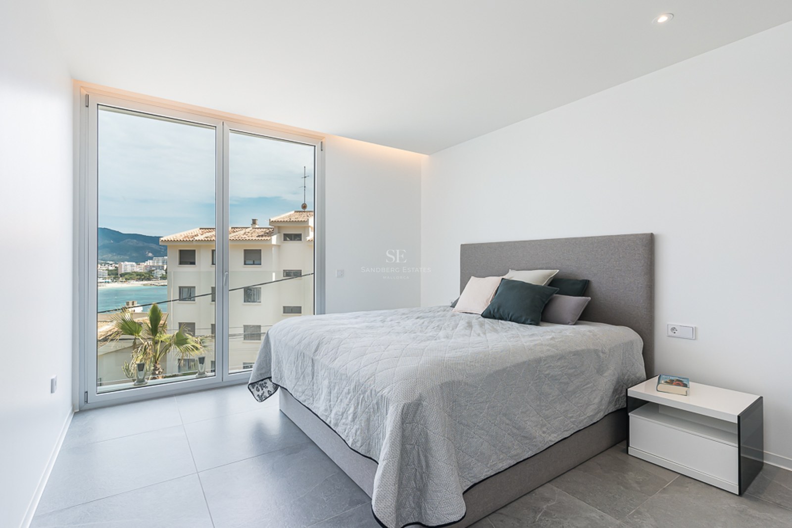 Bright modern bedroom with a large window overlooking a coastal town and sea, featuring a grey bed and tiled floor.