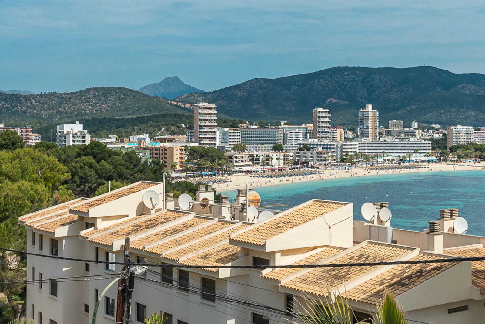 View overlooking a coastal bay with turquoise water, a sandy beach, white hotels, and green mountains in the background.
