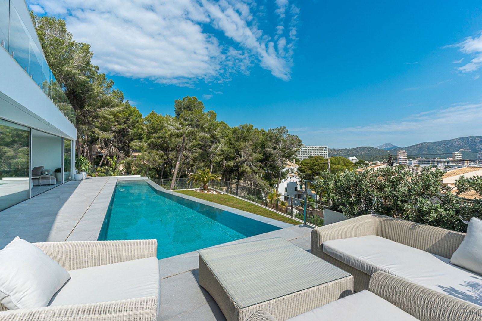 Modern terrace with a long blue swimming pool, white wicker lounge furniture, glass railings, and forest views.