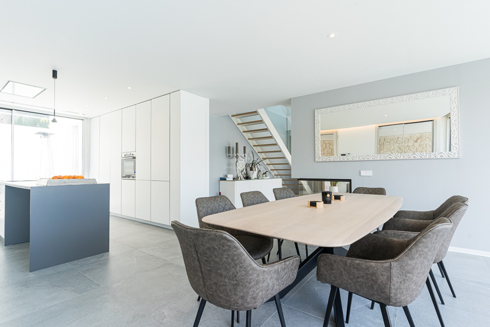 Bright dining area featuring a light wood table, grey chairs, and a minimalist white kitchen in the background.