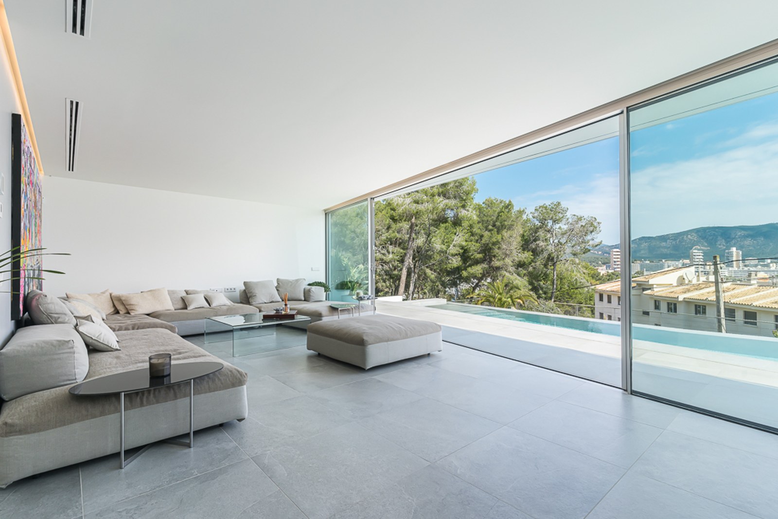 Spacious minimalist living room with grey sectional sofas and floor-to-ceiling glass doors overlooking a pool and mountains.