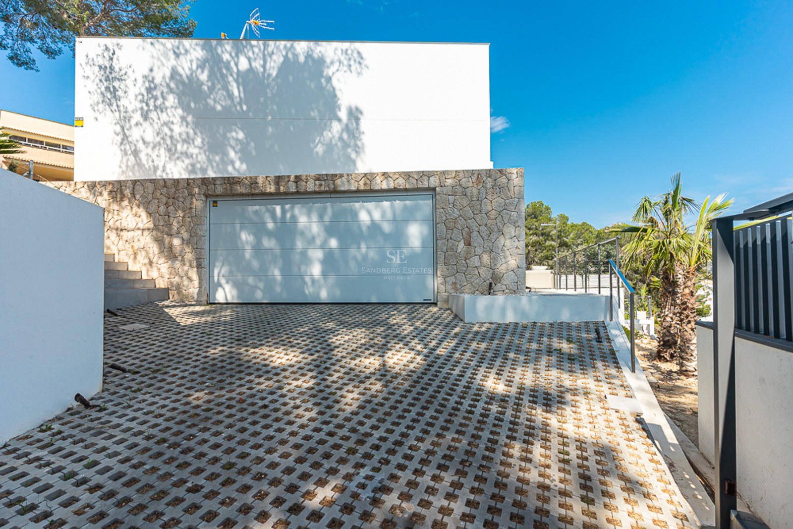 Modern white garage door in a natural stone wall with patterned pavers and palm trees.